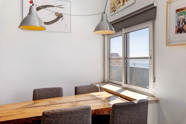 Un coin repas avec une table en bois, quatre chaises, deux lampes suspendues, une fenêtre donnant sur un paysage agréable et des œuvres d’art accrochées aux murs.
