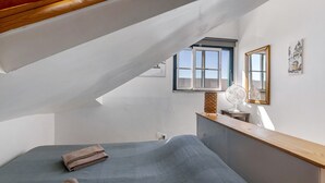 A bedroom with a sloped ceiling, a window, a bed, a bedside table, a mirror, and a fan.