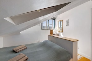 Une chambre avec un plafond en pente, un lit avec un matelas gris, deux serviettes pliées, une table de chevet avec une lampe et une fenêtre donnant sur un beau paysage.