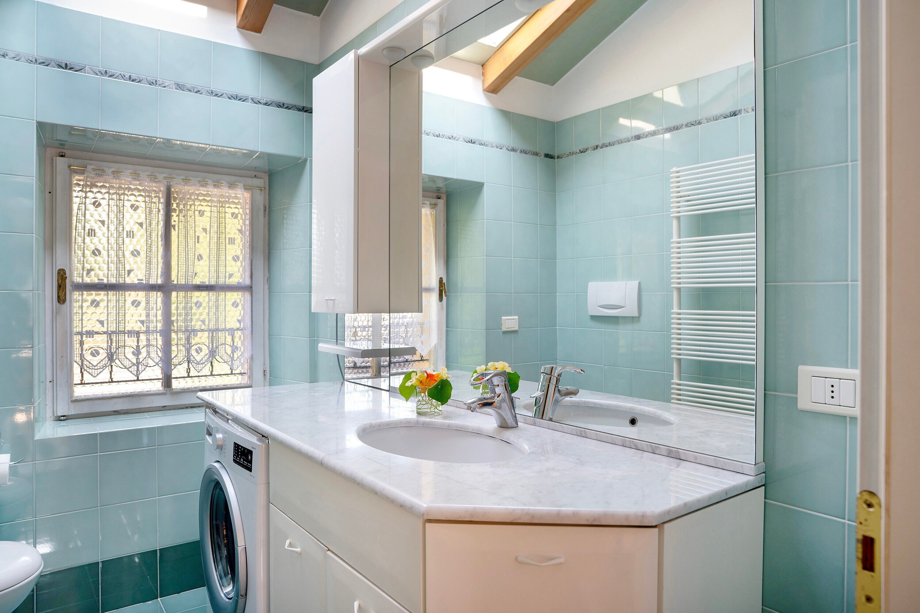 Een badkamer met een wasmachine, een wastafel, een spiegel en een raam met decoratief traliewerk.