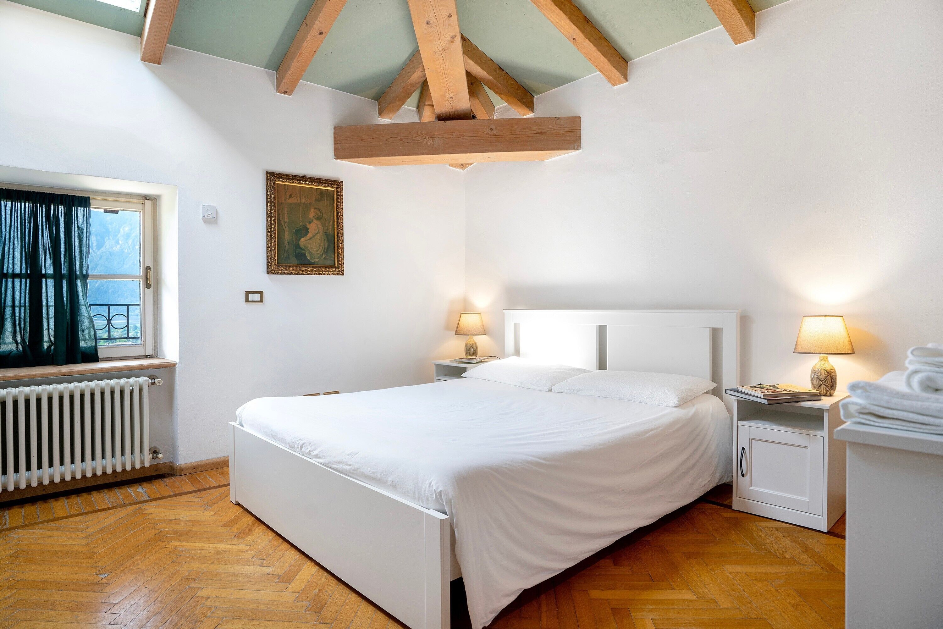 Une chambre à coucher avec un lit, des lampes de chevet, un radiateur, une fenêtre et un tableau accroché au mur.