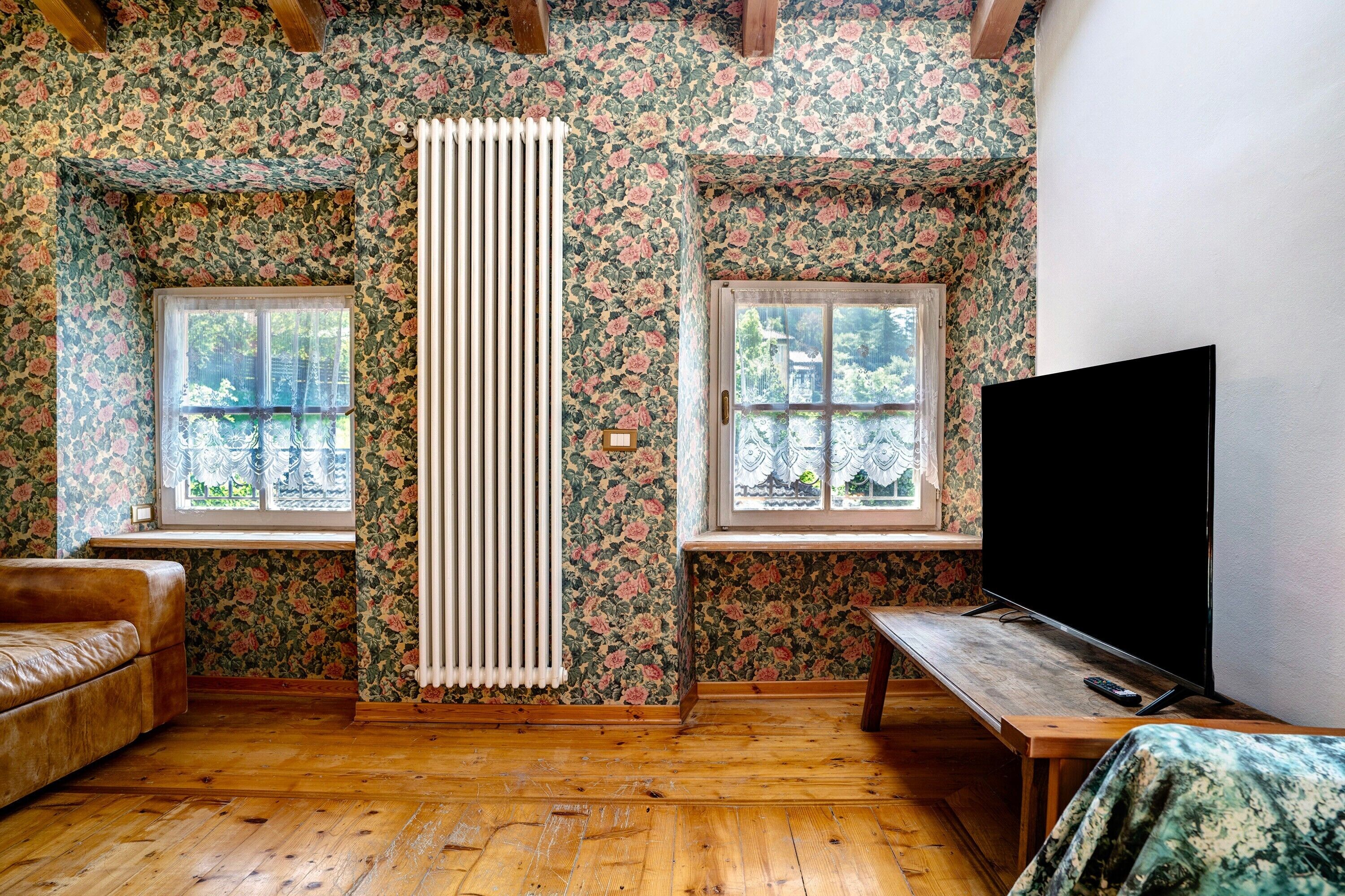 A room with floral wallpaper, a radiator, a wooden bench, a TV, and two windows.