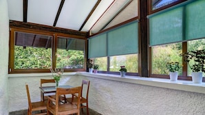 A covered patio with a wooden table and chairs, potted plants, and large windows overlooking greenery.