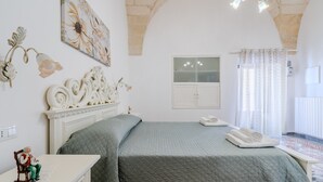 Ein Schlafzimmer mit Bett, Schminktisch, Nachttisch, fest installiertem Telefon, Fenster mit Vorhängen und einem Wandbild.