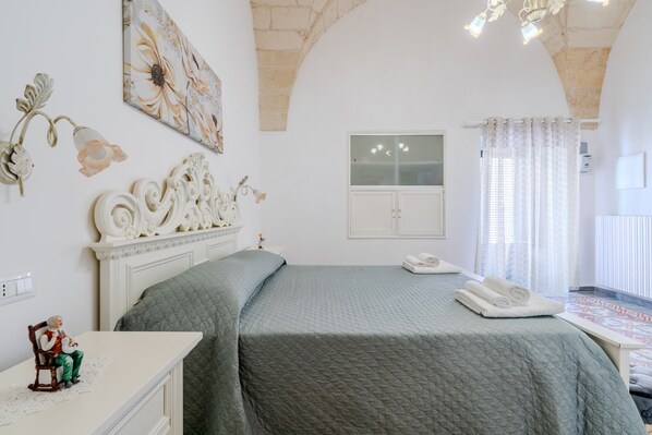 A bedroom with a bed, a dresser, a nightstand, a wall-mounted telephone, a window with curtains, and a decorative wall piece.