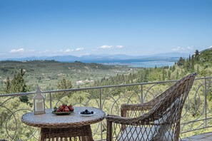 Een balkon met rieten meubilair, een kleine tafel met fruit en een kaars, en een prachtig uitzicht op heuvels en water.