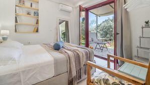 Un dormitorio con cama, una mesita de noche, estante con libros, espejo y vistas al exterior.
