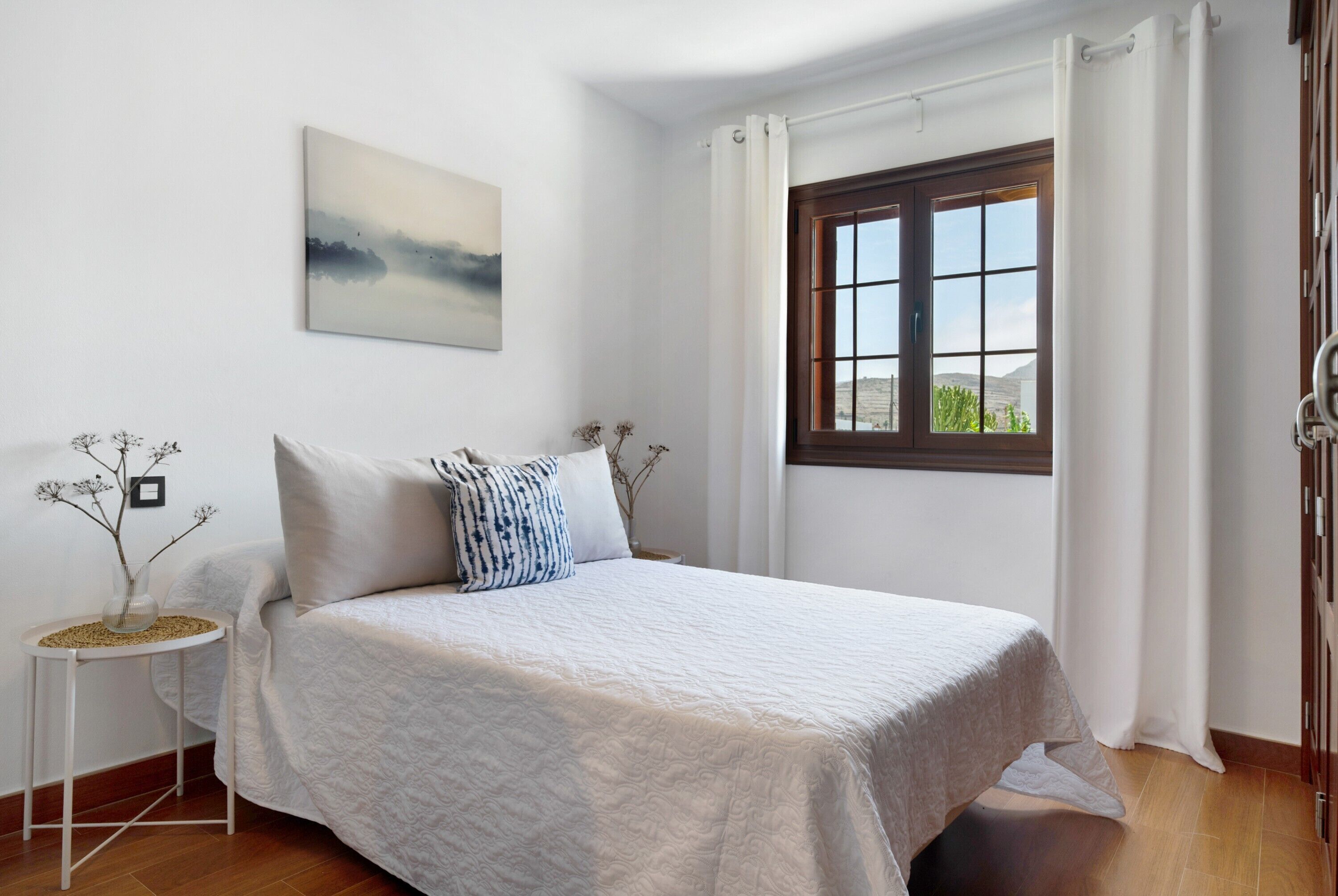 Un dormitorio con cama, ventana con cortinas, mesita de noche con un florero y un cuadro colgado en la pared.