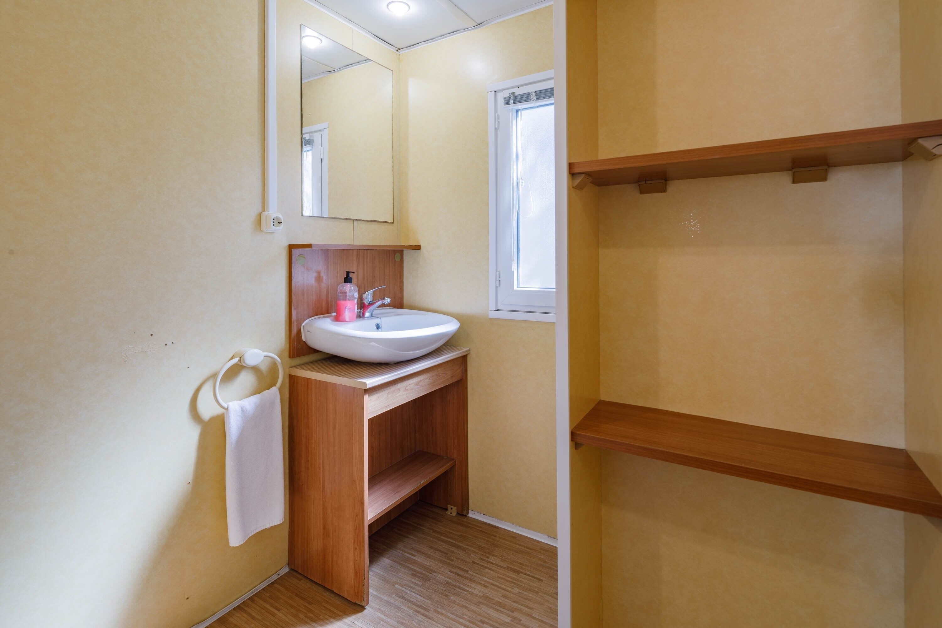 Un baño con lavamanos blanco, tocador de madera, un espejo, un aro para toallas y una ventana.
