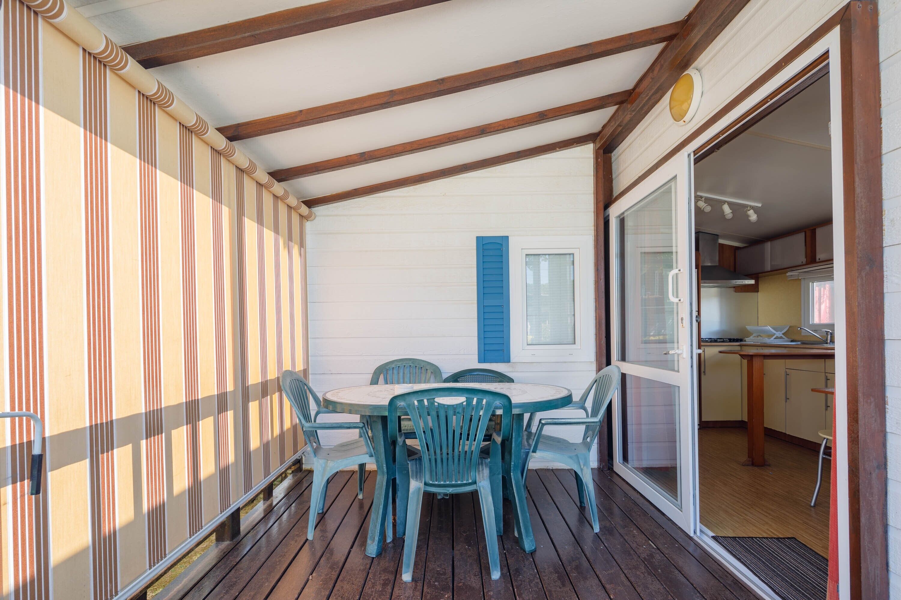 Un espacio al aire libre techado con una mesa y sillas, una puerta corrediza de vidrio hacia una cocina, y una pared a rayas.