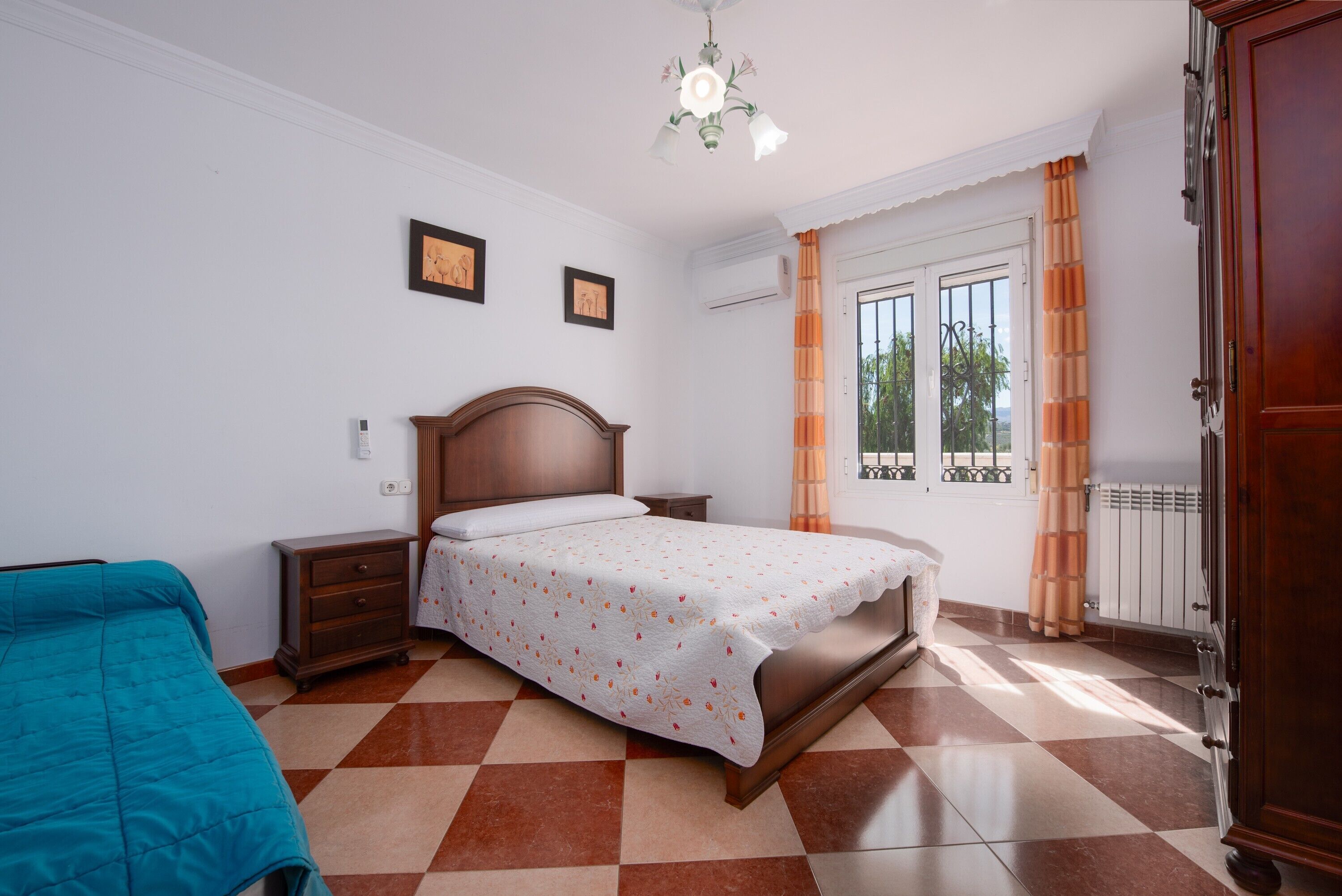 Une chambre avec un sol à carreaux, un lit recouvert d’une couvre-lit à motifs floraux, une table de chevet, une commode et une fenêtre avec des rideaux.