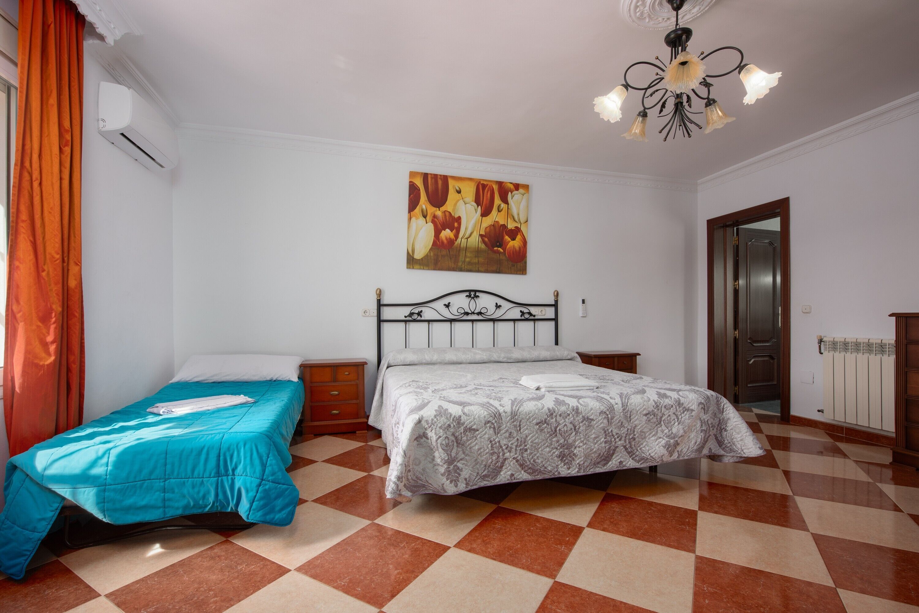 A bedroom with a checkered floor, a bed with a patterned bedspread, a nightstand, an air conditioning unit, a door, and a wall-mounted painting.
