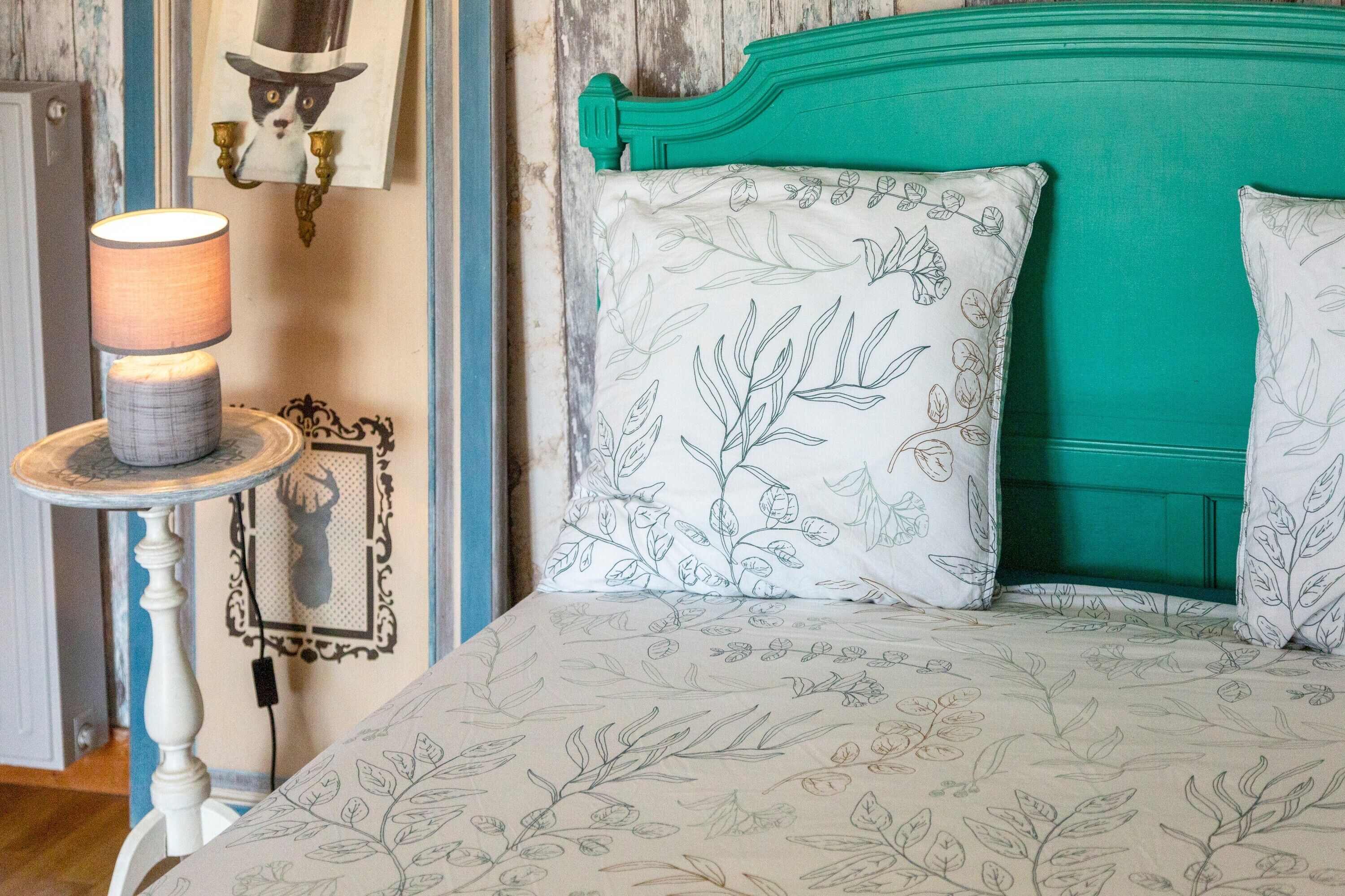 Uma cama com roupa de cama estampada com flores, cabeceira verde e luminária fixada na parede.