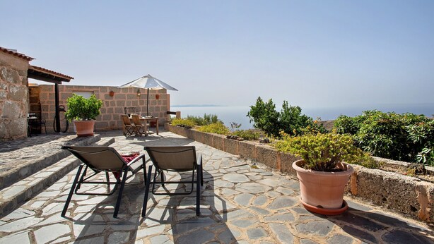 Eine Terrasse im Freien mit Steinboden, zwei Stühlen, einem Tisch, einem Blumentopf und Blick auf das Meer.