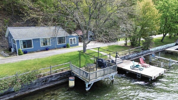 A house with a dock extending into a body of water, a grassy yard, and a tree in the background.