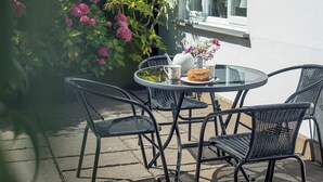 Une table ronde en verre, entourée de quatre chaises en osier noir, une théière blanche, deux tasses ornées de motifs floraux et une part de gâteau sur une assiette blanche.