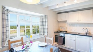 Une cuisine avec des meubles blancs, une cuisinière et un évier. On y trouve une table à manger recouverte d’une nappe bleue, ainsi que des chaises. Une fenêtre avec des rideaux laisse entrevoir la verdure à l’extérieur.