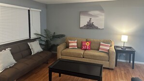 Un salon avec un canapé marron, une table basse noire, un canapé beige orné de coussins sur lesquels est écrit le mot « HOME », une plante en pot et un tableau accroché au mur.