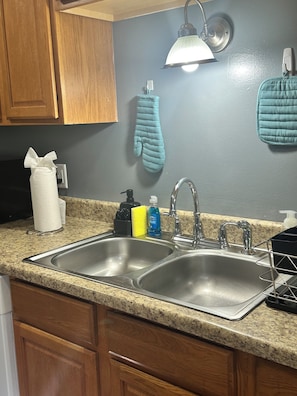 Una cocina con dos lavamanos de acero inoxidable, armarios de madera, encimera de granito, una lámpara colgante y un guante de cocina azul en la pared.