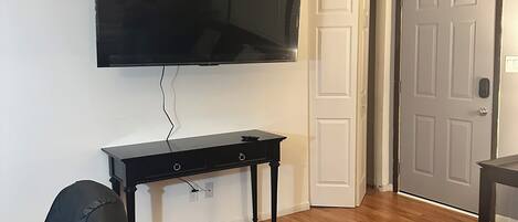 A living room with a flat-screen TV mounted on the wall, a black leather chair, a wooden floor, and a closed door.