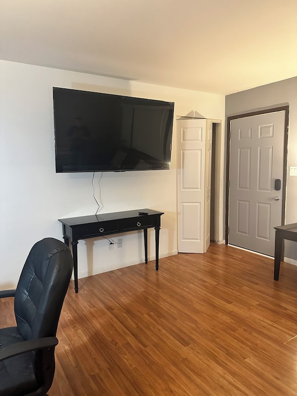 Una sala de estar con un televisor de pantalla plana montado en la pared, un sillón de cuero negro, piso de madera y una puerta cerrada.
