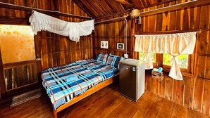 A wooden cabin room with a bed, a small refrigerator, and a window with curtains.