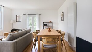 A modern living room with a wooden dining table, grey sofa, and wicker chairs.