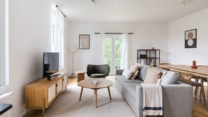 A modern living room with a grey sofa, a wooden coffee table, a TV on a wooden stand, and a dining area with a wooden table and chairs.