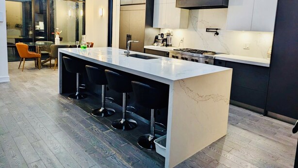 A modern kitchen with a central island, bar stools, a dining area with chairs, and a large window.