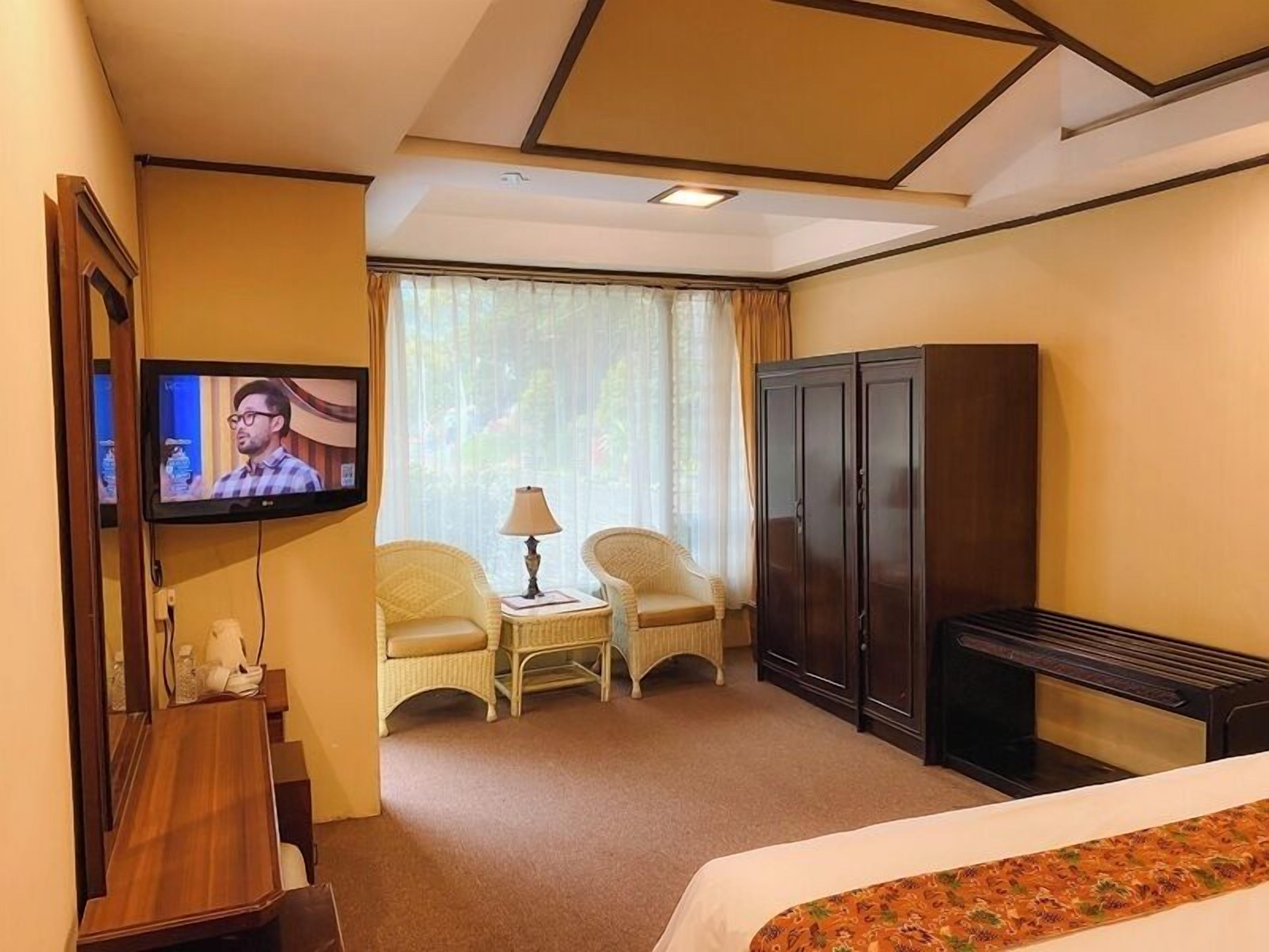 Una habitación de hotel con televisión, dos sillas, una mesita con lámpara, armario y cama.