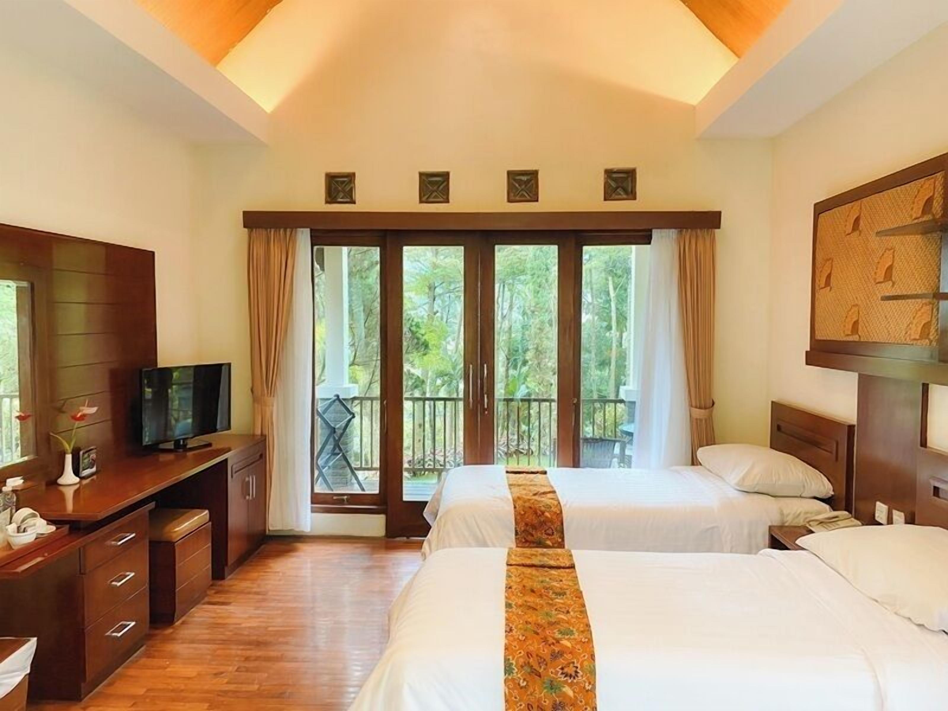 Habitación de hotel con dos camas, un escritorio con televisor y un balcón con vistas a zonas verdes.