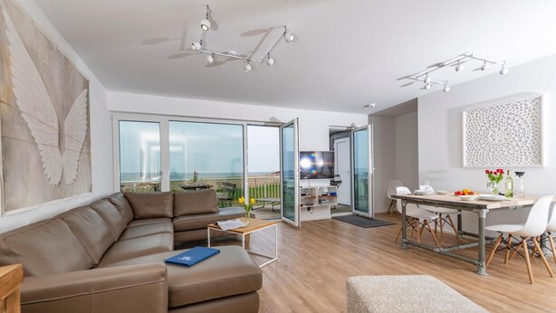 Een moderne woonkamer met een bank, een eettafel en een televisie. Er is een balkon met uitzicht op het strand.