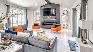 A modern living room with a fireplace, two orange armchairs, a grey sofa, and a ceiling fan.