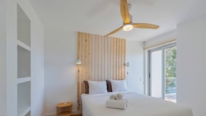 A modern bedroom with a wooden slat headboard, a large bed with white linens, a ceiling fan, and a view of the outdoors through a sliding door.