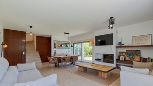 A modern living room with a white sofa, a dining area with a wooden table and chairs, a fireplace, and a TV mounted on the wall.