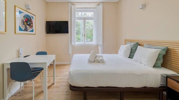 A bedroom with a bed, a desk, a chair, a TV, and a window.