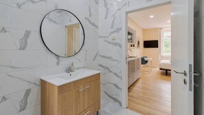 A modern bathroom with marble walls, a round mirror, a wooden vanity, and a view into a living area with a sofa and TV.