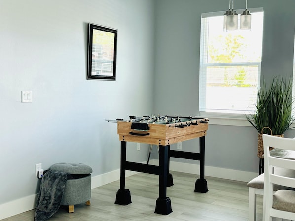 A room with a foosball table, a small ottoman, a chair, a window with blinds, and a potted plant. - Brand New Tampa Oasis with Private Jacuzzi Retreat (Lutz)