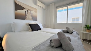 A hotel room with two beds, a painting on the wall, an air conditioning unit, a window with curtains, and a small table with a plant. - AP408 ACAPULCO PLAYA SM NERJA (Nerja)