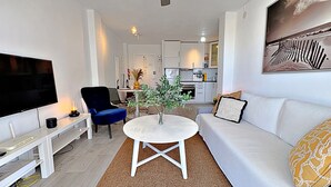 A modern living room with a flat-screen TV, a blue chair, a white sofa, a coffee table, and a kitchen in the background. - AP408 ACAPULCO PLAYA SM NERJA (Nerja)