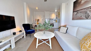 A modern living room with a flat-screen TV, a blue chair, a white sofa, a coffee table, and a kitchen in the background. - AP408 ACAPULCO PLAYA SM NERJA (Nerja)
