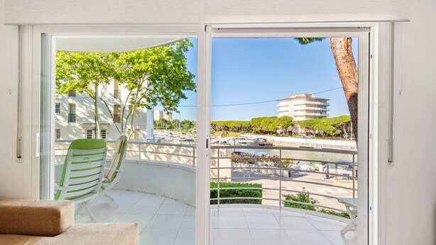 A balcony with a green-striped chair, a beige sofa, and a view of a building and marina. - WELCS APARTAMENTO 170 PDA en el puerto (Platja d´Aro)