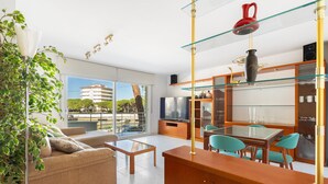 A modern living room with a sofa, coffee table, and dining area with chairs. There is a large window with a view of buildings and trees. - WELCS APARTAMENTO 170 PDA en el puerto (Platja d´Aro)