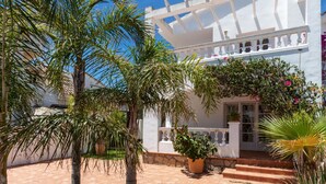 A white building with a balcony, surrounded by palm trees and potted plants.