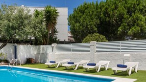 A swimming pool with white lounge chairs and blue cushions, surrounded by a white wall and greenery.
