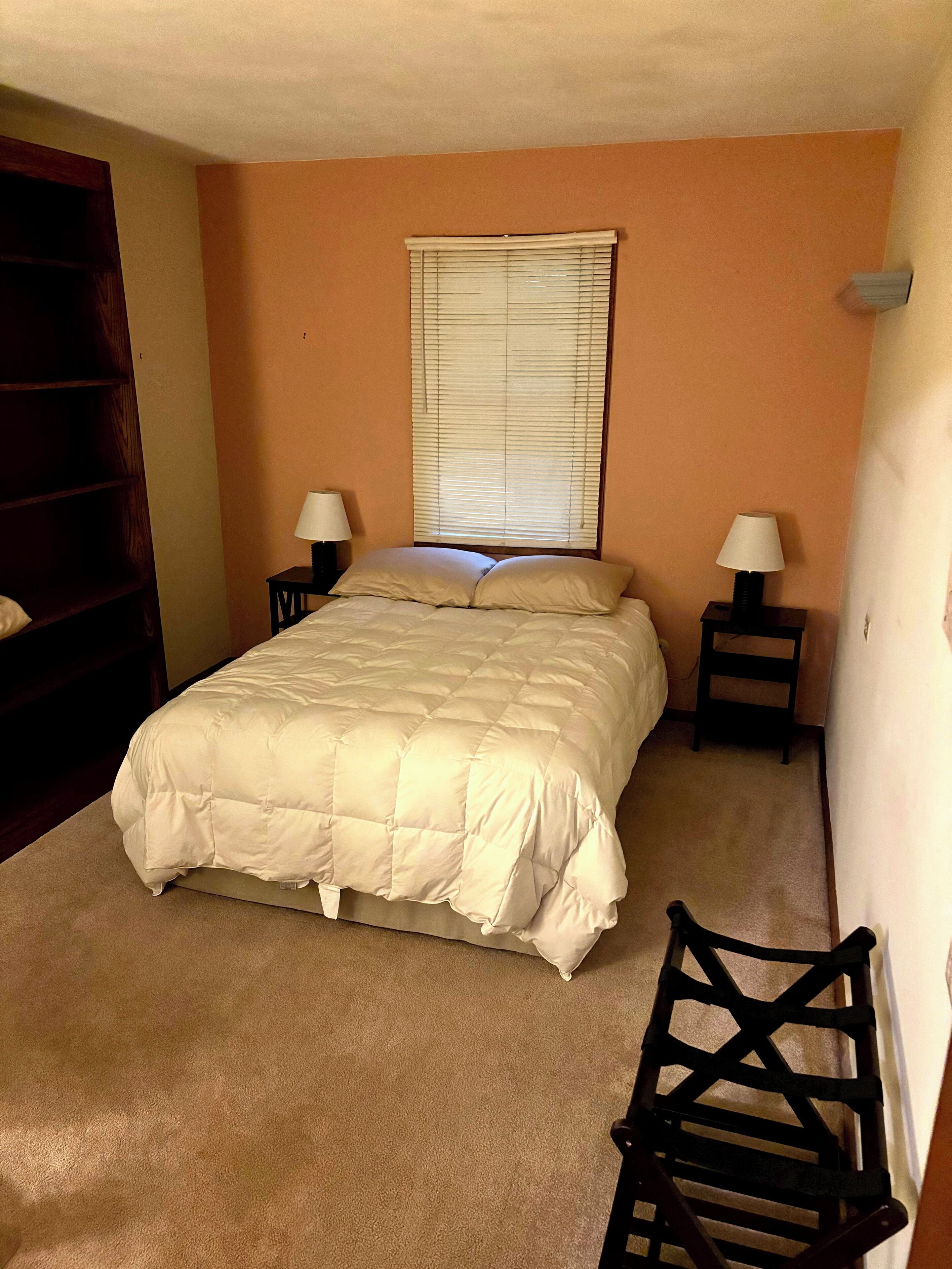 A bedroom with a bed, two bedside tables with lamps, a window with blinds, a chair, and a wooden wardrobe.