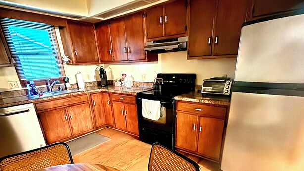 A compact kitchen with wooden cabinets, a stainless steel refrigerator, a stove, and a dining table with chairs. - Family Friendly 4 Bedroom House Near Penn State University (State College)
