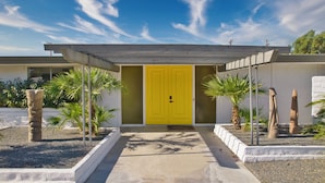 A modern house with a yellow door, a concrete pathway, and palm trees.