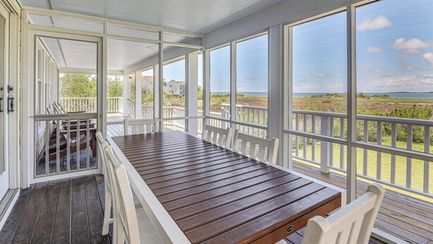 Eine überdachte Terrasse im Freien mit Holztisch und weißen Stühlen, große Fenster mit Blick auf Strand und Meer.