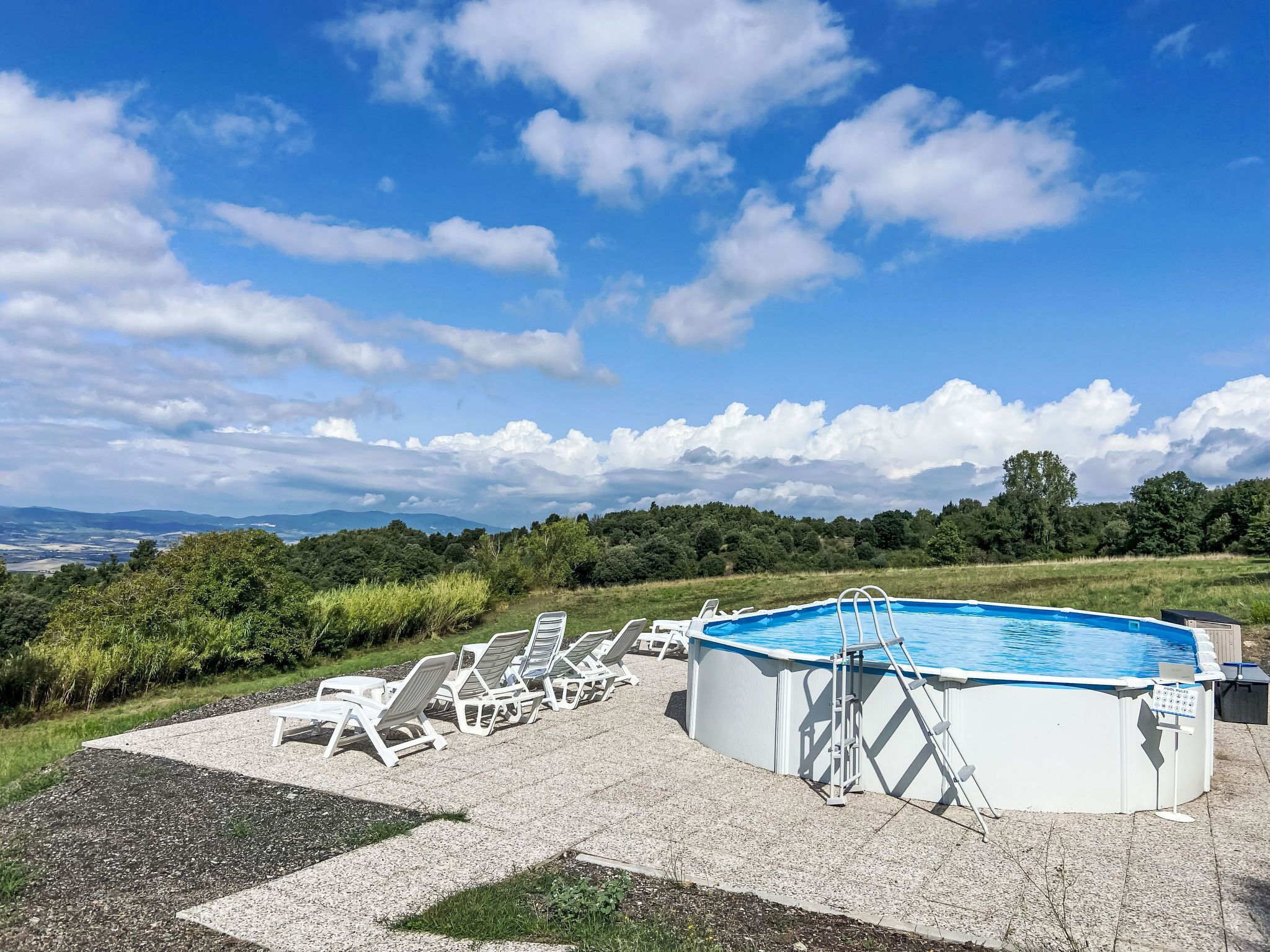 A swimming pool with a ladder, surrounded by white lounge chairs, set against a backdrop of a clear sky and distant hills.